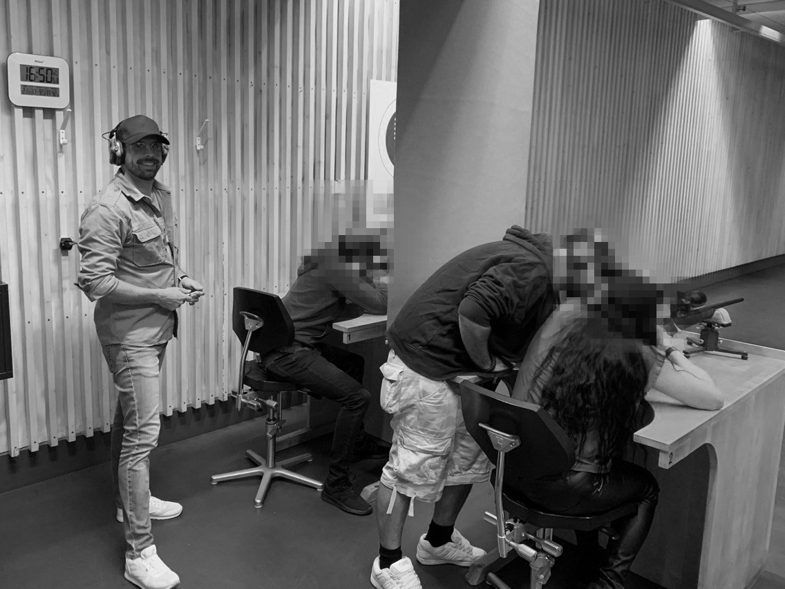 Four people at an indoor shooting range; one standing wearing ear protection and cap, others seated at shooting benches.